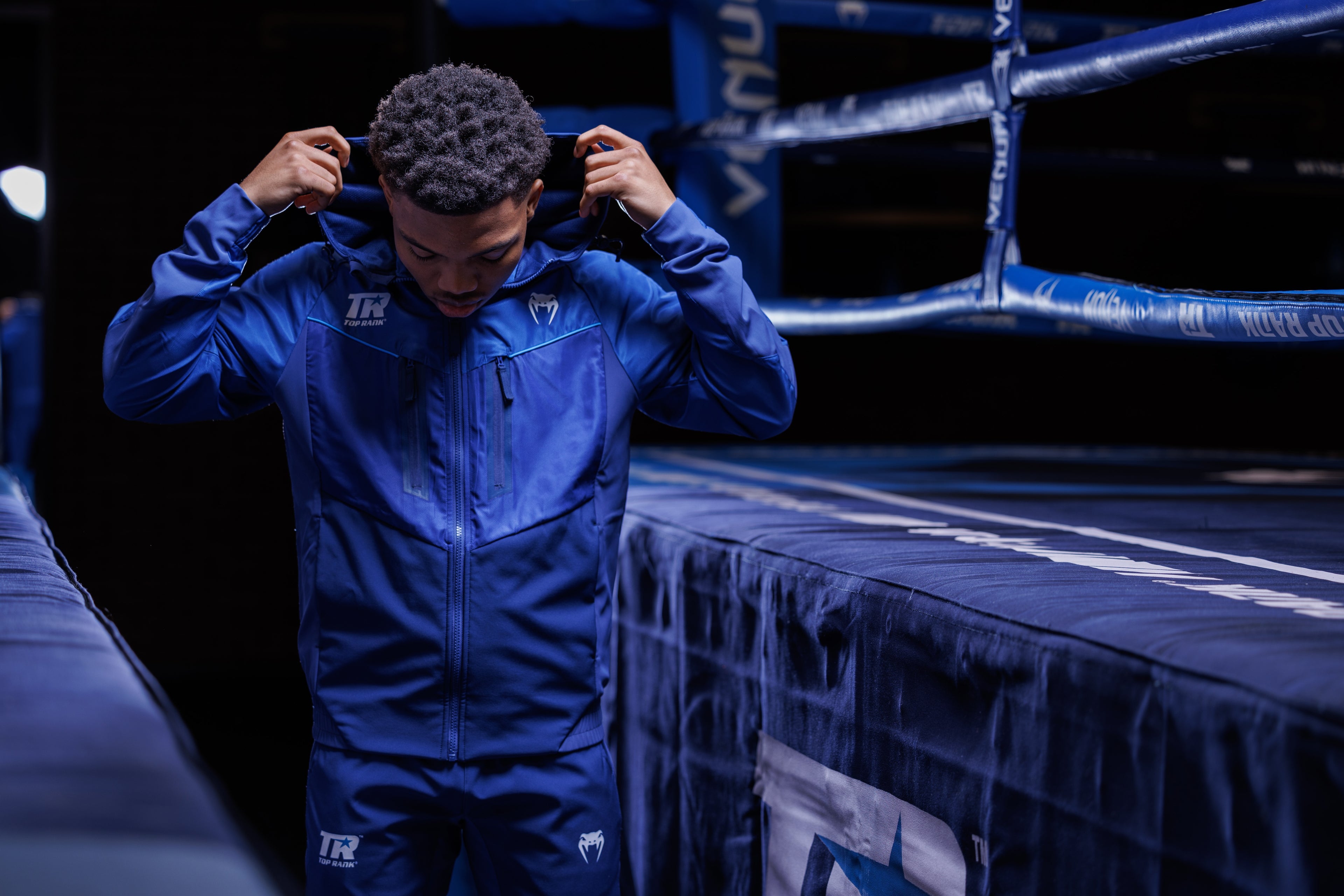 A boxer in a blue tracksuit with 'Top Rank' and Venum logos, adjusting the hood while standing next to a boxing ring with blue ropes and padding, captured in a dimly lit setting