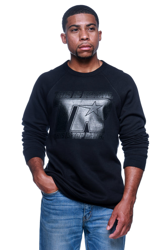 Person wearing a black crewneck sweatshirt with a black-on-black TR logo with gel imprint on the front chest, paired with blue jeans.